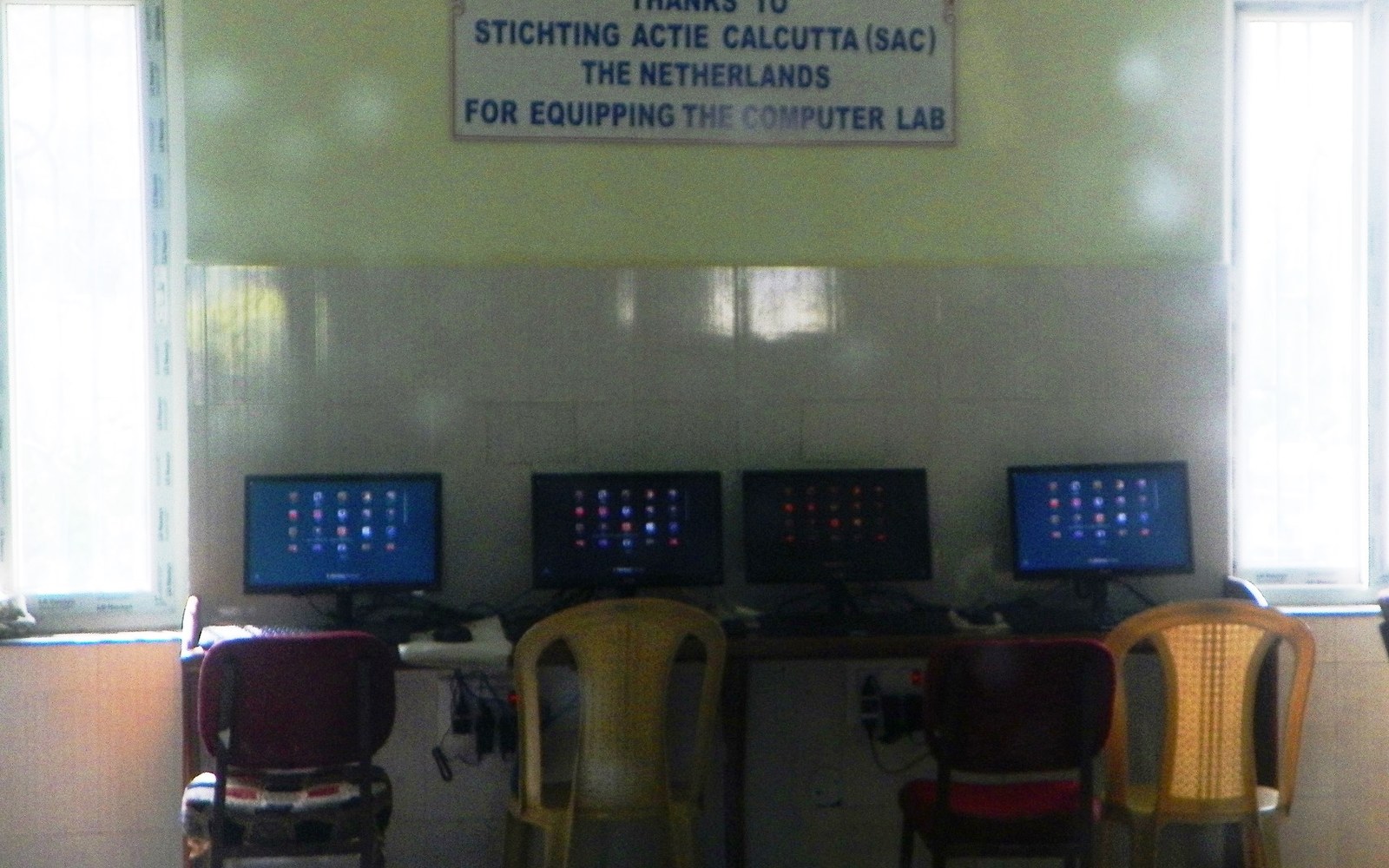 Four computer monitors on a desk below a sign thanking Stichting Actie Calcutta for the lab